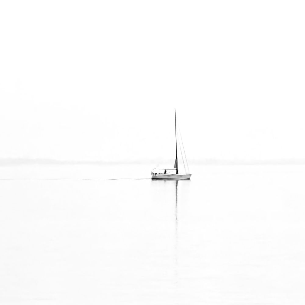 SW Aufnahme eines kleinen Segelboots ohne gehisste Segel. Das Boot ist nicht mehr ganz mittig im Bild. Der Mast spiegelt sich im ruhigen Wasser. Der Rest des Fotos ist nur weiß, bis auf eine leicht transparente graue Linie am Horizont. 