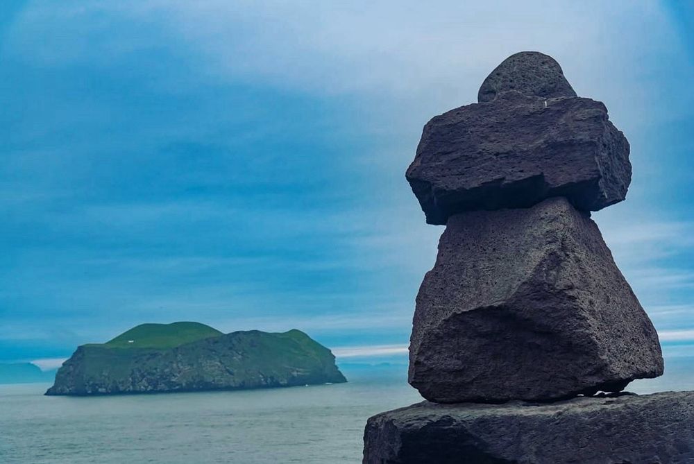A standing stone sculpture with four rocks balanced atop one another sits in the foreground with the ocean, a cloudy blue sky; and a smaller island in the background.