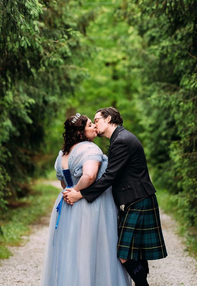 A photo of a man and woman kissing in an aisle of fir trees. She wears a blue dress laced with blue ribbons in the back and a crystal crown. He wears a black, green, and yellow striped kilt and a wool jacket. They hold hands behind her back as she tilts her face up into the kiss.