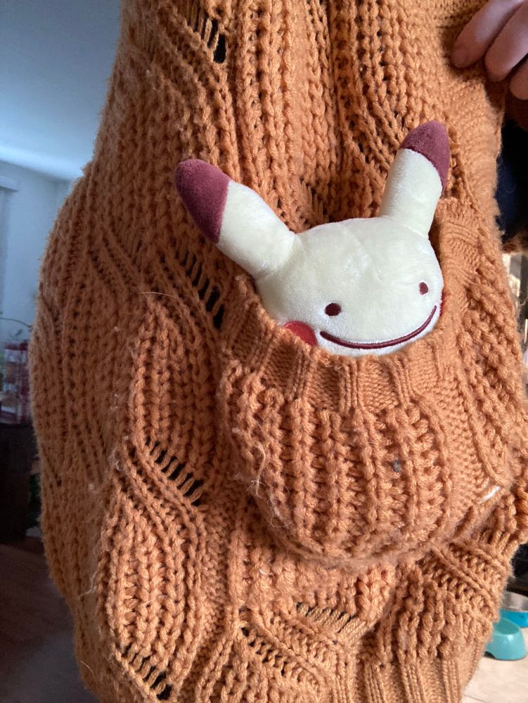Photo of a Pokemon plushie in a dark orange cardigan. The plushy is a Ditto as a Pikachu, and only its face smile and up are visible from the pocket. 