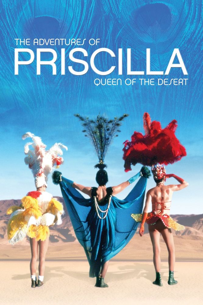 The adventures of Priscilla Queen of the Desert image shows a desert landscape with three people backs to viewer - from left fethery white and yellow with exposed legs, peacock themed headress with flowing blue cape attached to hands held out to the sides, mostly nude with a large plume of red on head and a red/yellow tutu.