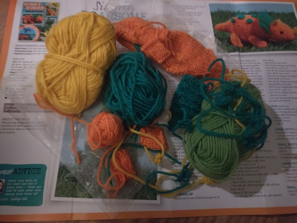 A little pile of wool, in green, blue, yellow, and orange, including the components elements of a knitted triceratops 
