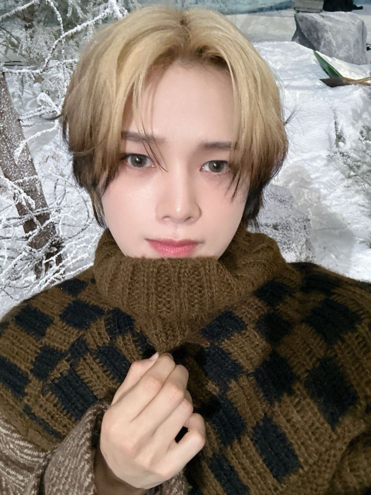 K from &Team on the set of the Yukiakari MV. He's wearing a brown and black knitted jumper that has a huge rolled collar almost up to his mouth. His hair is blond with dark tips and he's staring straight at the camera. 