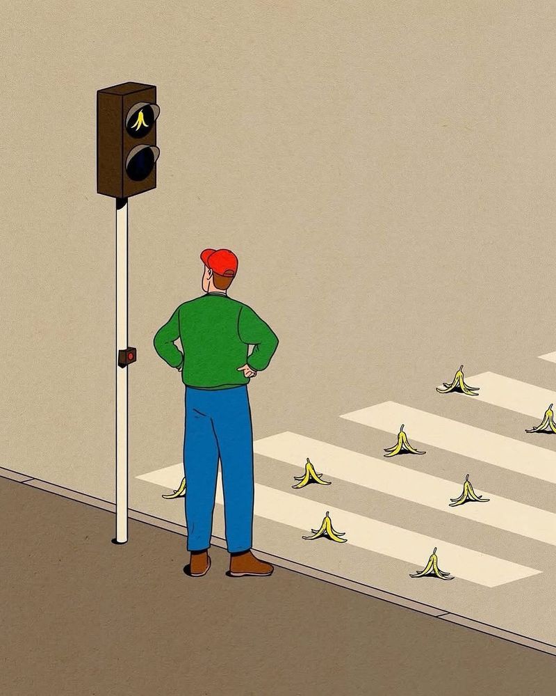 A man stands on the corner, waiting for the light to change. The crosswalk is strewn with banana peels. The light is neither red or green, it is on banana.