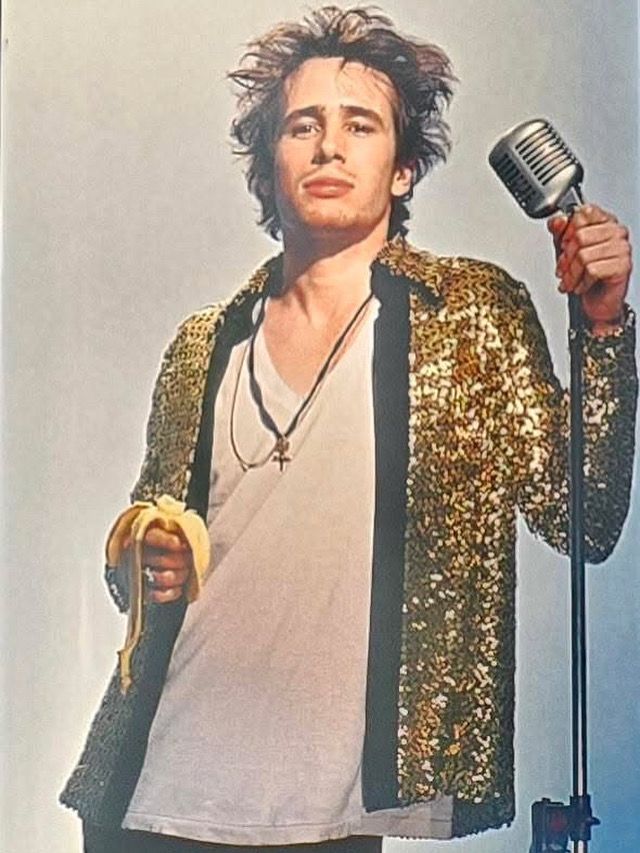 Photograph of Jeff Buckley, wearing a gold sequin jacket and a white T-shirt, holding a microphone in one hand in a banana and the other.