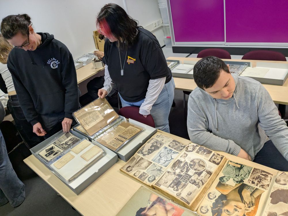 Two students are standing up, going through the pages of an album with photographs and other papers. A third student is sitting next to them looking at a scrapbook 