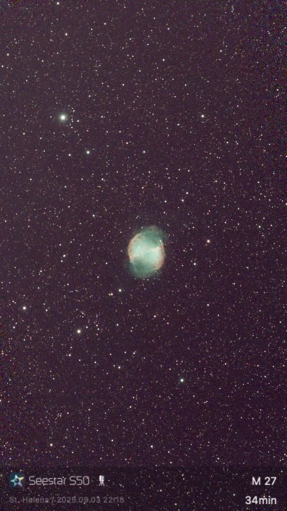 M27, the Dumbell Nebula, close multicolored against a starry background.  In this image space isn't dark, because the waxing moon is giving a reddish-brown hue.  The nebula itself looks a bit like an apple core turned sideways in the image.  There are two rims of redish at eiter side.  Whitish green stretches a between the rims, narrowing towards where they meet.  Green puffs spill out the top and bottom.