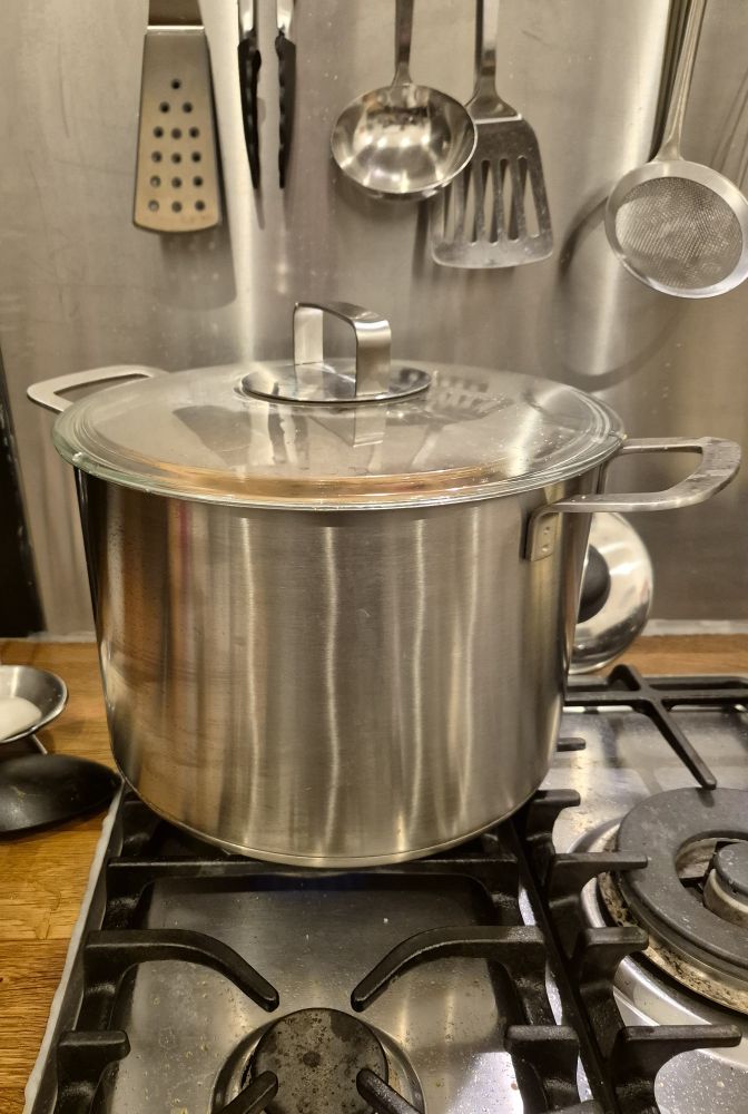 Silberner Topf mit Deckel auf einem Gasherd. Im Hintergrund hängen Küchenutensilien.