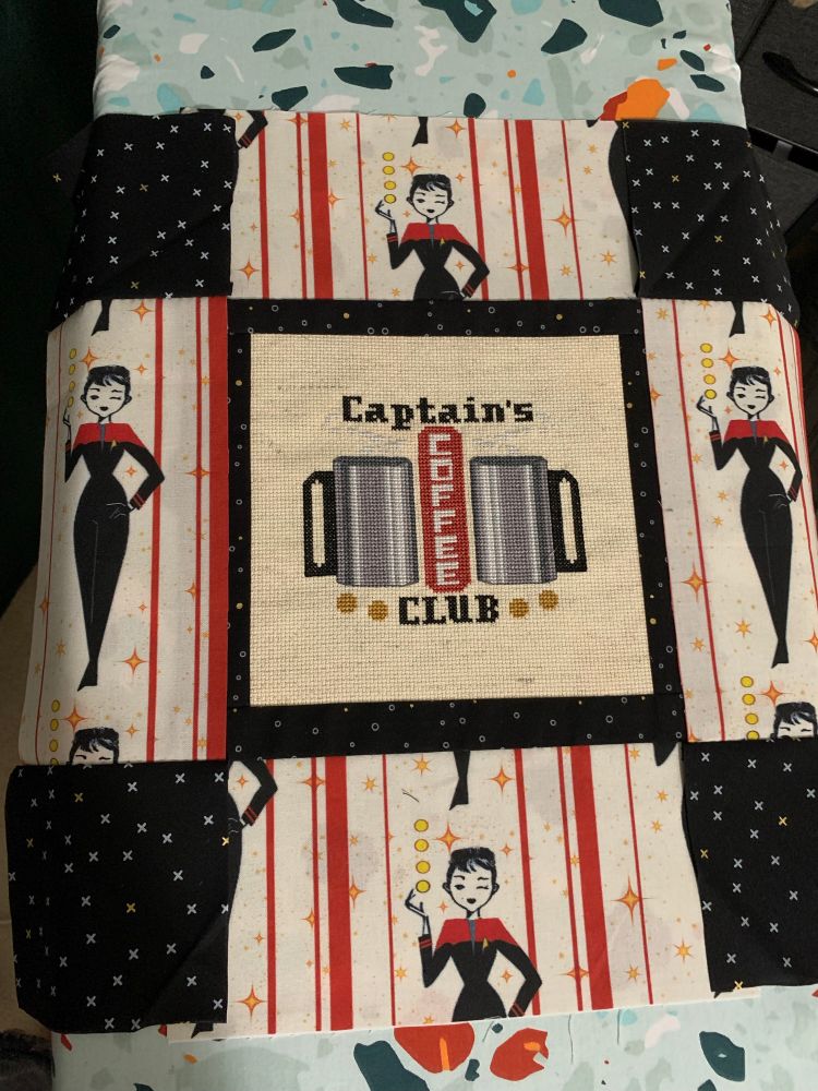 Picture of a cross stitch with fabric sewn onto the sides. The cross stitch says, “captain’s coffee club” and has two stainless steel coffee mugs. There are two types of fabric, black in a small border around the cross stitch and then at the corners and another fabric which has red stripes and a stylized figure of a woman in a Starfleet uniform, meant to be captain Janeway, but the face doesn’t really look like her. 