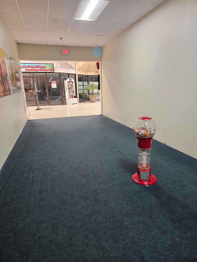 Horror movie ass hallway in a completely empty mall, with a single gumball machine dropped sorta in the middle, but just off center enough to be truly twisted, and also(not pictured) the gumballs all look like they're from 1995, and in the store next to this they're selling t-shirts that say "Guns are like boobs, even the small ones are fun to play with," and importantly these are the least offensive shirts on display