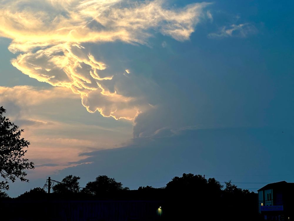 A large storm cloud partially lit from behind and below by the setting sun. Protuberances of cloud hang from under the anvil. Most of the rest of the cloud is in deep shadow, and no detail can be seen.
