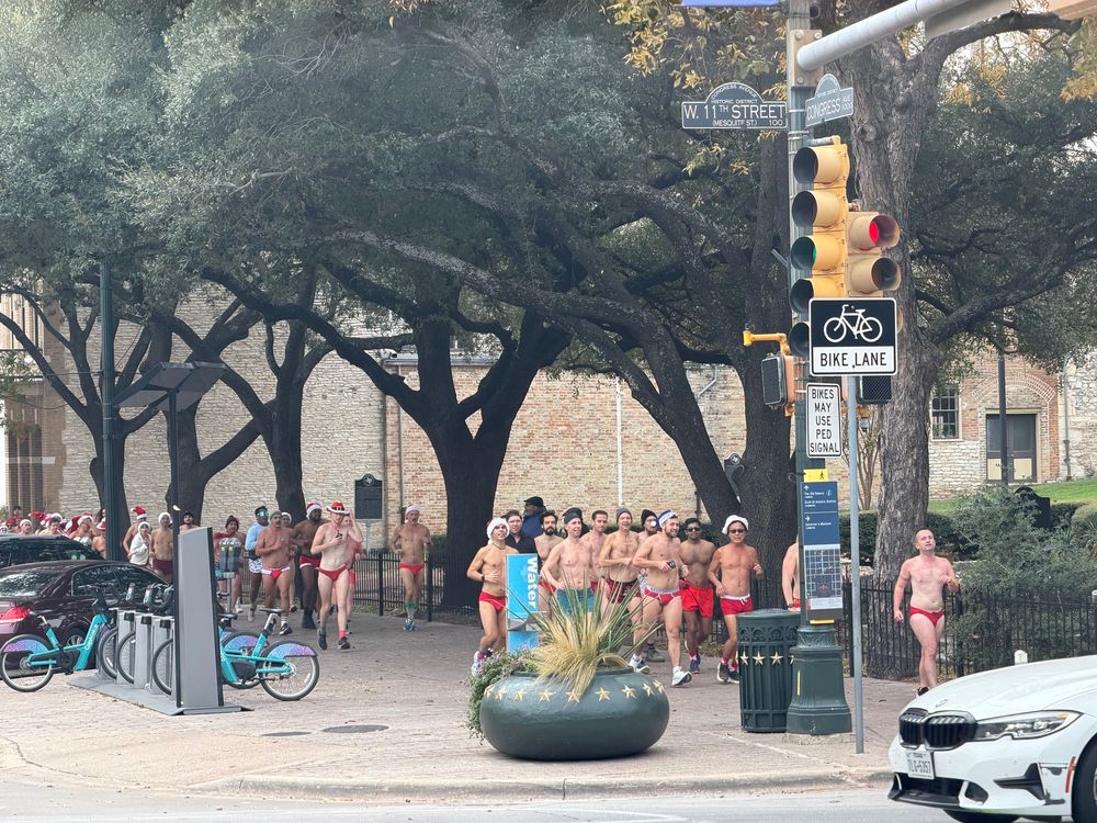 A group of men, mostly in red briefs or Speedos, running down a sidewalk in an urban area.