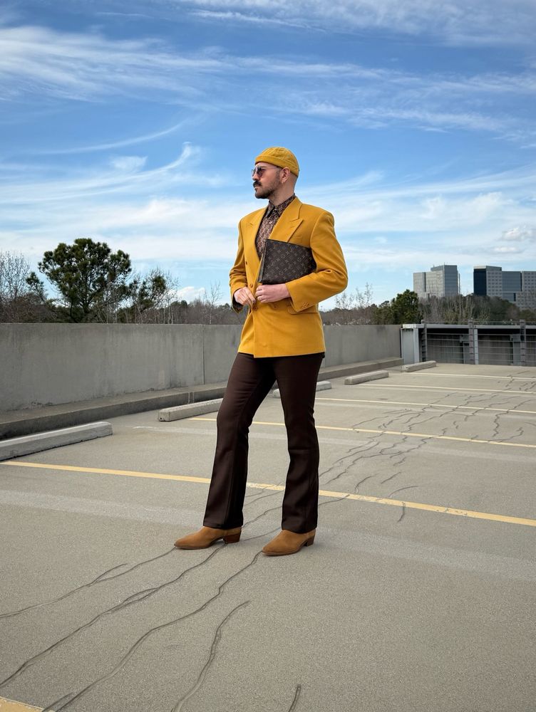 Standing at a parking lot wearing brown thrifted vintage Calvin Klein blouse and a mustard blazer    pared with brown Wrangler pants, light brown Celine boots, round Balenciaga sunglasses and monogram Louis Vuitton clutch. 