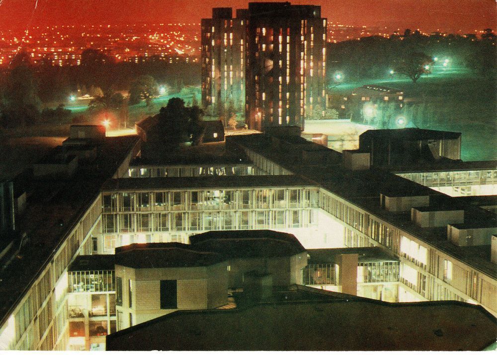 A colour picture postcard showing 1960s concrete buildings, arranged in quadrangles, lit up ; there are tower blocks in the middle distance, and beyond that trees and parkland, and then a distant town. The effect of the lighting is such that the buildings to the front are a creamy pale yellow; the parkland emerald and malachite, and the town beyond a pale crimson