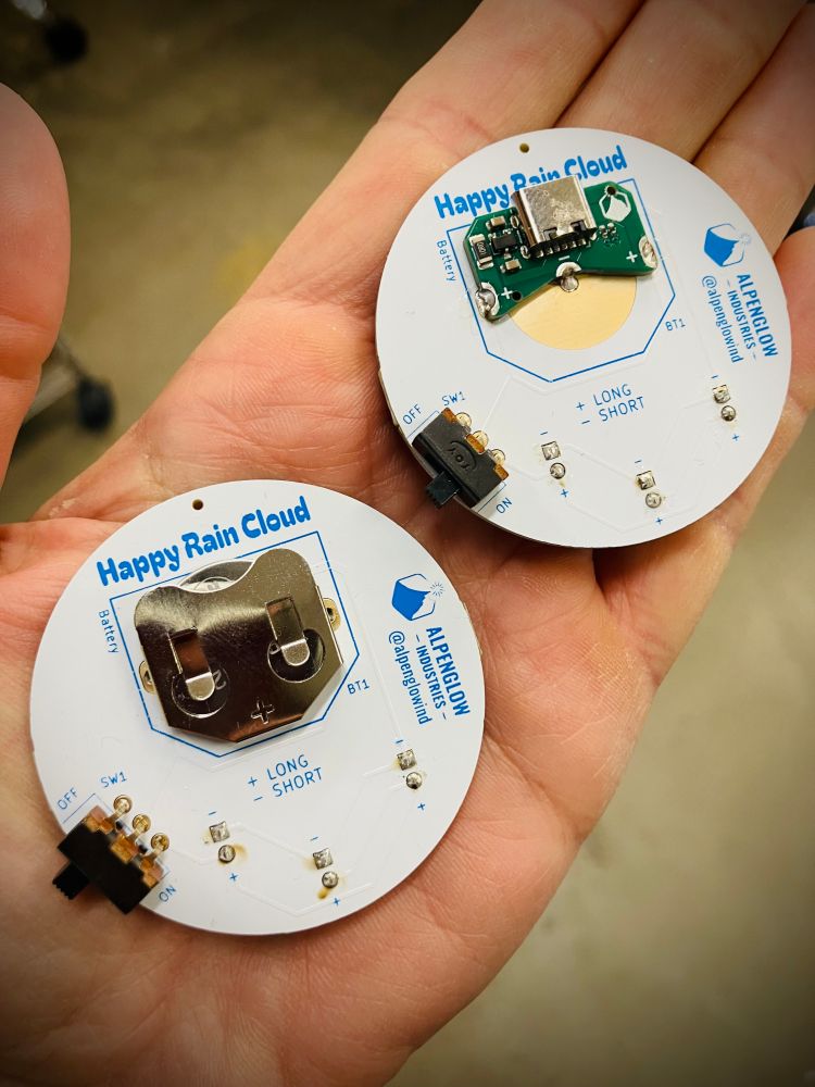 Two round circuit boards with white soldermask and blue silkscreen, left shows a CR2032 holder and the right shows a USB adapter board soldered into its place instead.