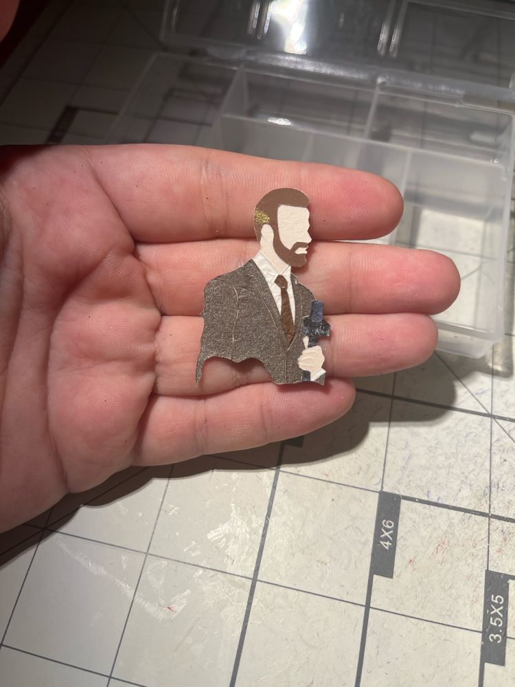 a very small papercraft of james hinchcliffe in a brown-gray suit holding a microphone. the papercraft is under 2 inches in height and is resting on a hand