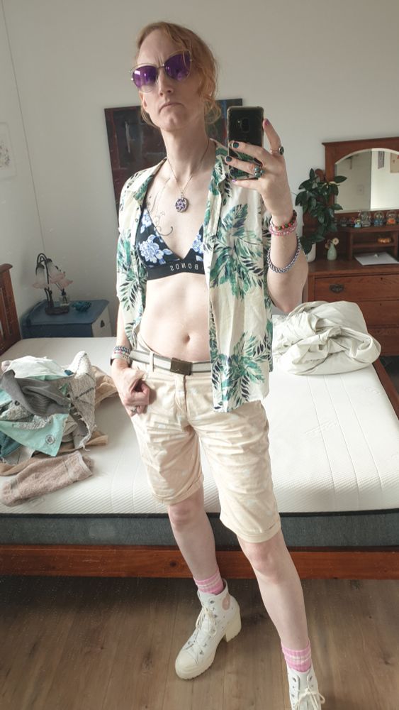 Mirror selfie in the guest bedroom of my test outfit a few days prior. Towels and bedclothes are dumped on the mattress.

I'm wearing an open shirt with vague palm designs on it. My open chest shows off my black/blue Bonds swim top. I have tight-ish shorts just above my knees, my white Converse heels, and large, purple sunglasses.