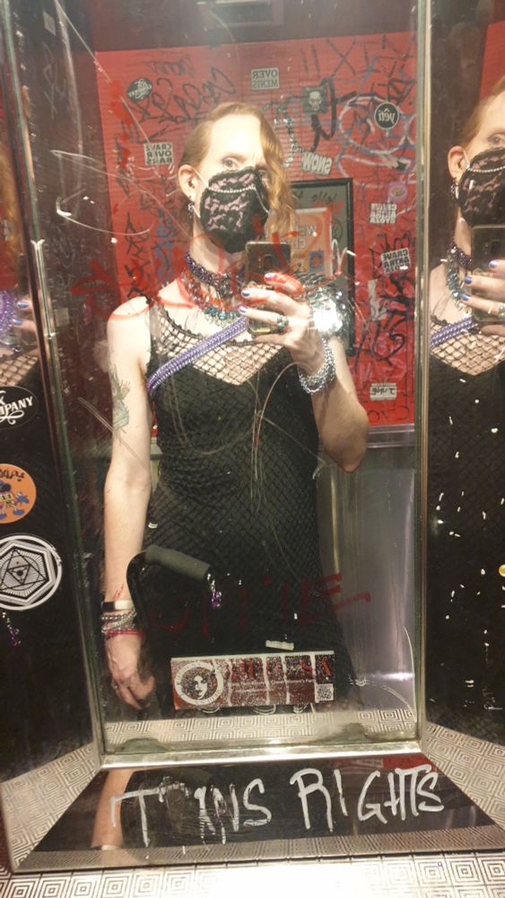 Mirror selfie in the bathroom at the event. Three mirrors with different angles make it look almost like a triptych.

I'm wearing my pink mask with lace covering and a chain. I'm holding my walking stick. My outfit is much more visible here.

There are stickers and graffiti on the mirror, and on the wall behind me. Prominently at the bottom of the mirror "TRANS RIGHTS" is scrawled in white. The atmosphere makes me look much cooler than I am.