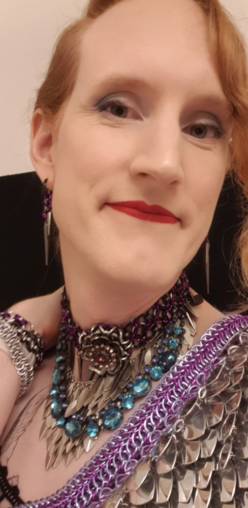 Close-up selfie that shows off a lot of my chain accessories. I'm wearing black/purple/grey spike earrings, three bracelets (silver, grey, black and purple), my very detailed purple and black necklace with hanging spikes and a drapey blue gem chain all layered on top of a second necklace of scale mail with either leaves or feathers. In the foreground part of my shoulder pauldron is showing, with a bit of detail from the strap showing, as well as the scales.

I'm also wearing makeup and smiling slightly. I like this look.