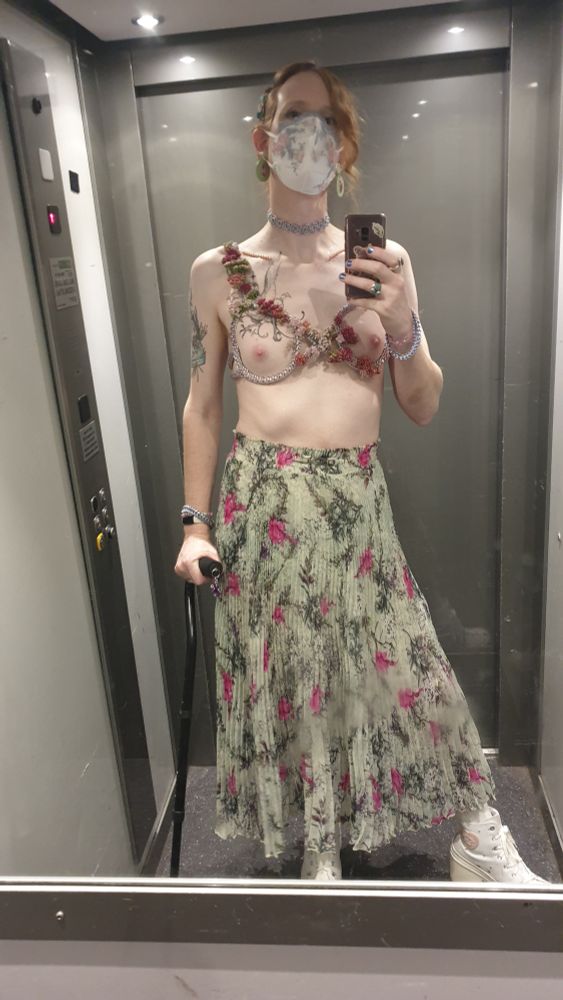 Elevator mirror selfie. I'm supporting myself slightly with my walking stick. I'm wearing an ankle-length, teal/mint skirt with dark pink flower and plant patterns on it. I'm wearing my cream-coloured converse heels.

On my top I'm only wearing my elastic, chainmaille bra, which has some flowers weaved into it from one shoulder to the bottom of the other side. My tits and nipples are bare, as the "bra" doesn't actually cover that area.

Sadly, my chest tattoo is covered a lot by the flowers. I thought this was the best side to weave them in because my hair is partly down on the other shoulder, but now I'm not sure.

I have a maille choker, some bracelets, and a white mask covered in light blue and pink lace.

I mostly like this outfit, but I wish the execution of flower-weaving throguh maille was done better, and there's something about the skirt that gives a good look but I feel I was after something else.