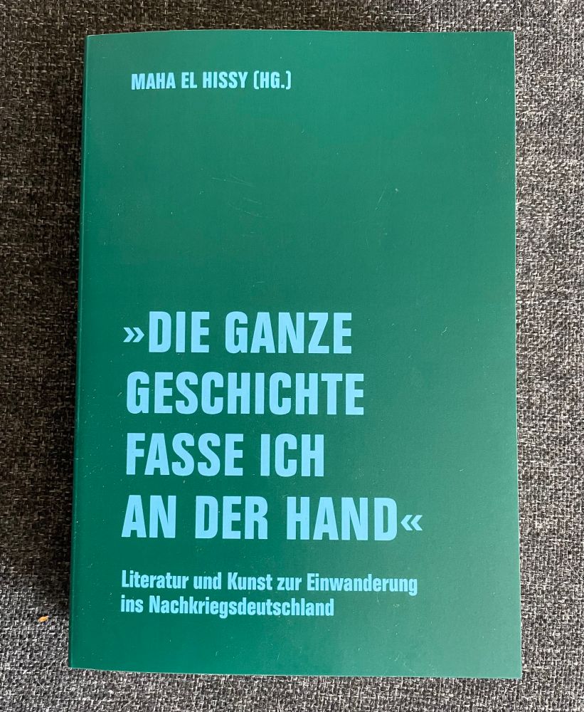 Das Buch, hellblaue Schrift auf grünem Grund, auf dunkelgrauem Stoff liegend.
