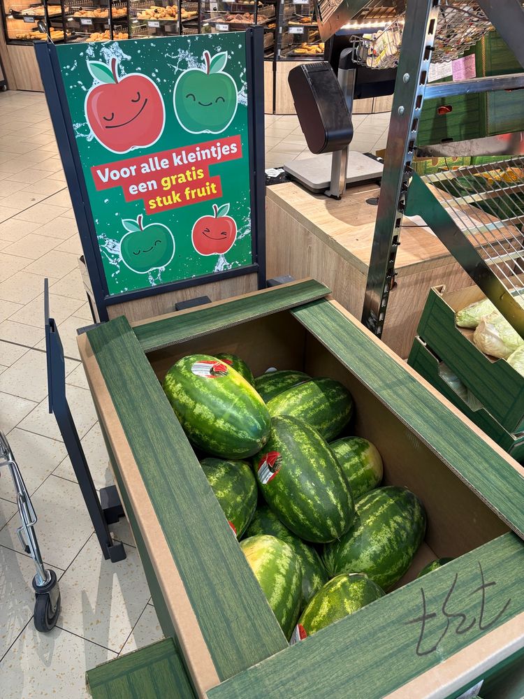 een bord met “Voor alle kleintjes een gratis stuk fruit” staat achter een bak met gigantische watermeloenen ter grote van een pasgeboren baby