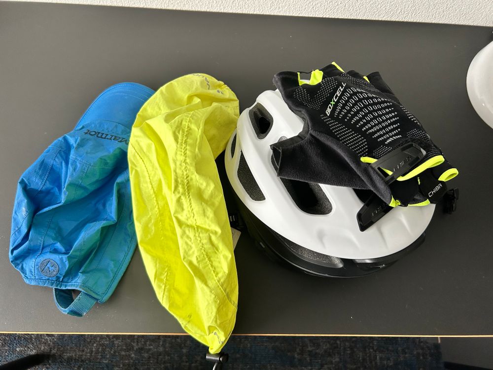 Neben dem weißen Helm und den Handschuhen liegt ein neongelber Regenüberzug für den Helm und eine regenabweisende Schirmmütze