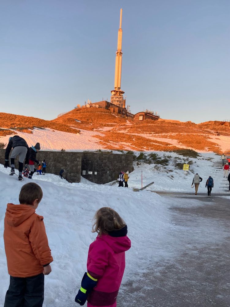 Un petit garçon et une petite fille enroulés dans des combi de ski au pied de l’antenne du Puy-de-Dôme 
Il y a de la neige au sol un ciel bleu parfait et une lumière rosée sur l’antenne blanche et le sommet du Puy 