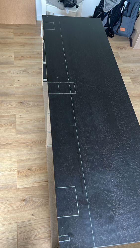 Lange schwarze Kommode mit weißen Markierungen auf der Oberfläche, umgeben von Holzfußboden und Rucksäcken im Hintergrund