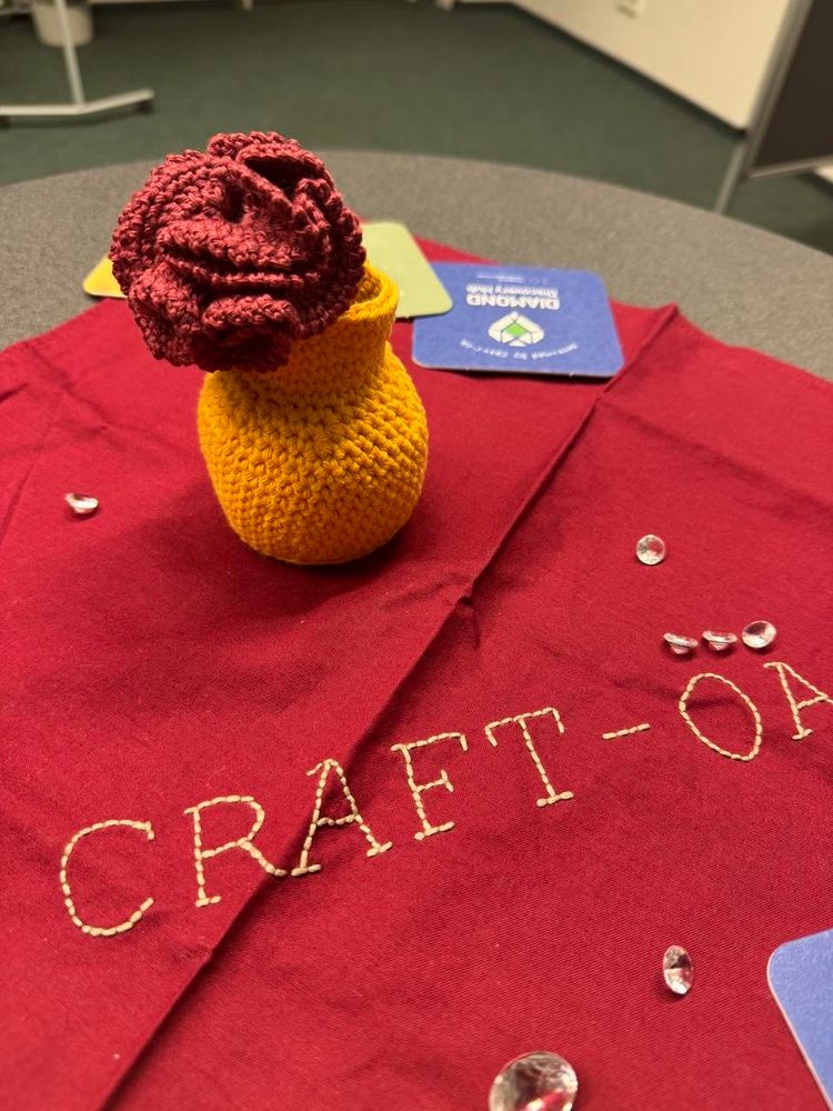 A red crocheted flower in a yellow crocheted small vase on a table covered with a red tablecloth with “Craft-OA” stitched on