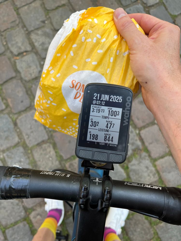 Blick vom oben auf Radcomputer, der Startzeit 7:12 und 100km anzeigt. Außerdem im Bild eine Hand die Brötchen-Tüte hält