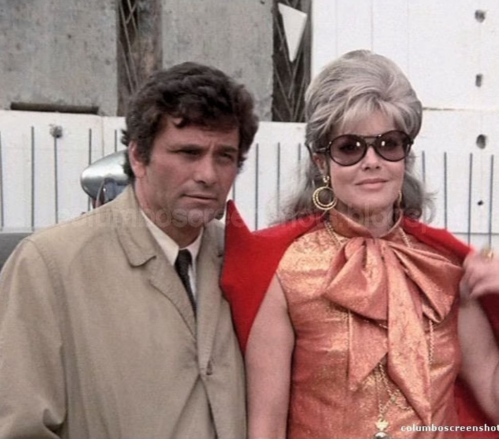 Peter Falk, as Columbo, wearing a tan trench coat stands with Janis Paige, as Goldie, wearing glamorous sun glasses and a bronze brocade outfit with a large neck bow.
