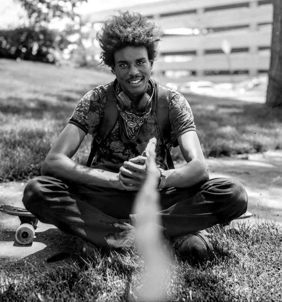 Black and white image of a teenager sitting on his skateboard.