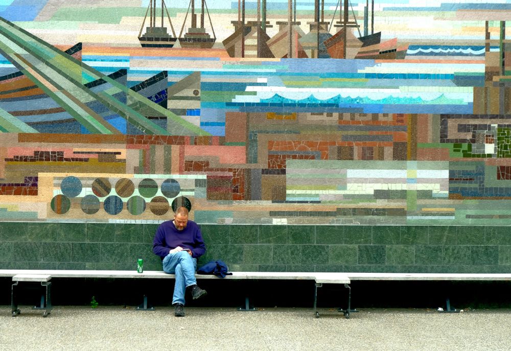 A man sitting on a bench, engrossed in his phone in front of a colorful mosaic mural.