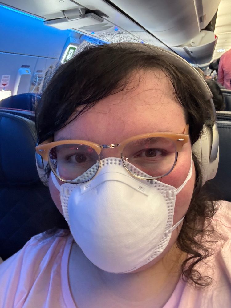 Picture of Brie wearing a mask and noise cancelling headphones on an airplane.
