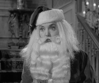 Carolyn Jones as Morticia Addams as Santa Claus. Black and white still of her with a massive white beard and a santa hat.