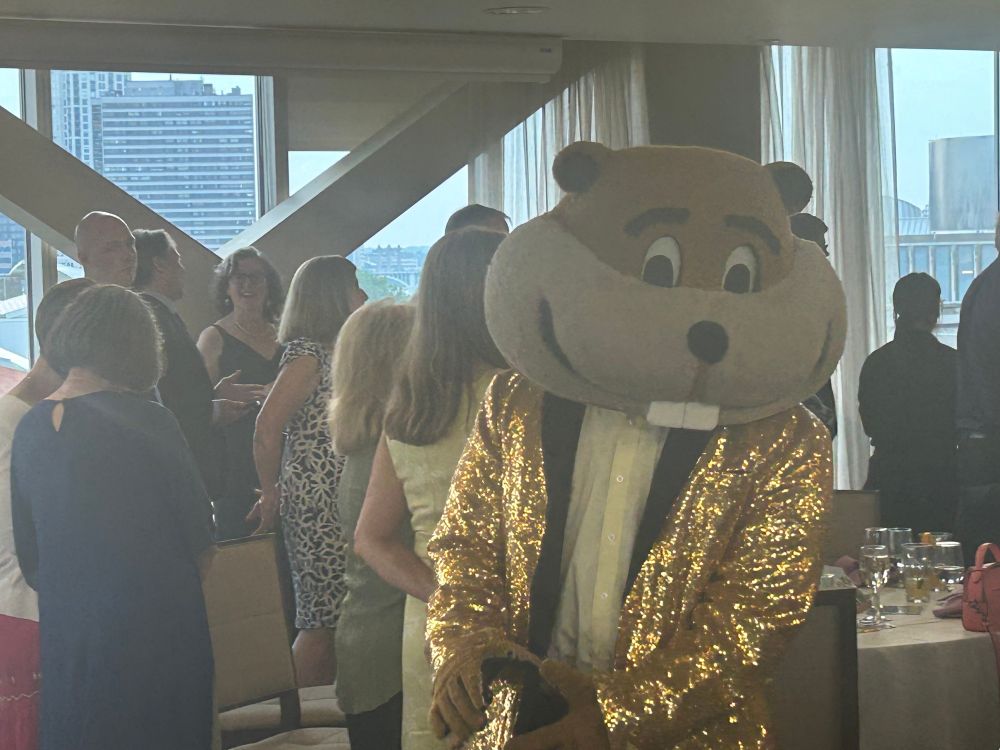 An enormous headed gopher mascot in a gold coat smiles impishly at the camera while everyone in the background watches something else (not pictured).