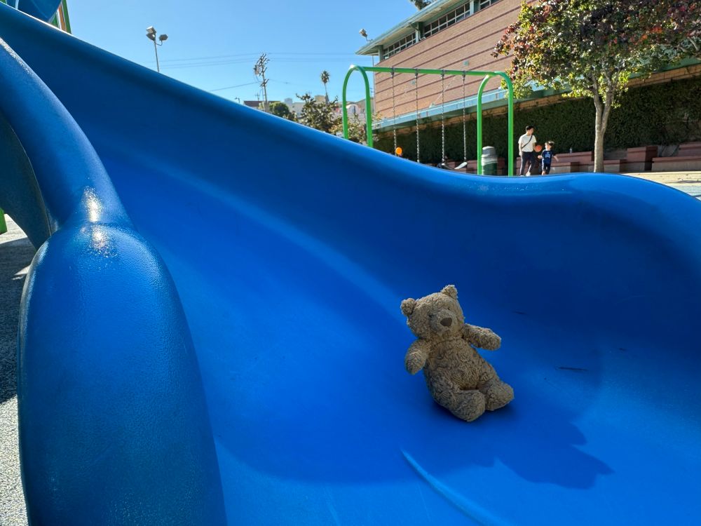 A small brown teddy bear at the bottom of a blue slide. 