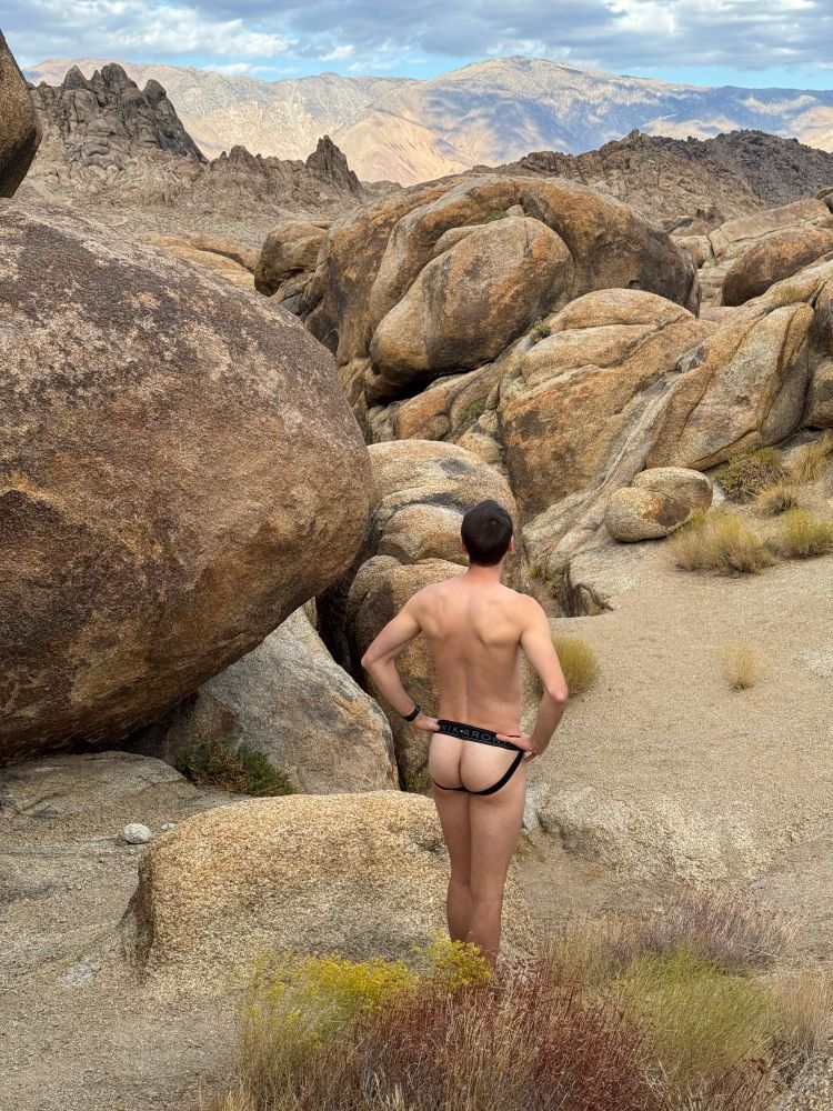 Man standing in rock field with a jockstrap on facing away from camera 
