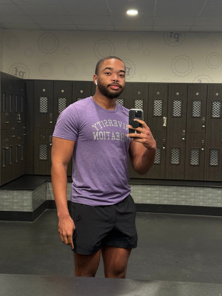 Mirror selfie of me in a light purple shirt that’s tight around my underarm and chest area and 4 inch black shorts. I’m looking in the mirror as I snap the pic.
