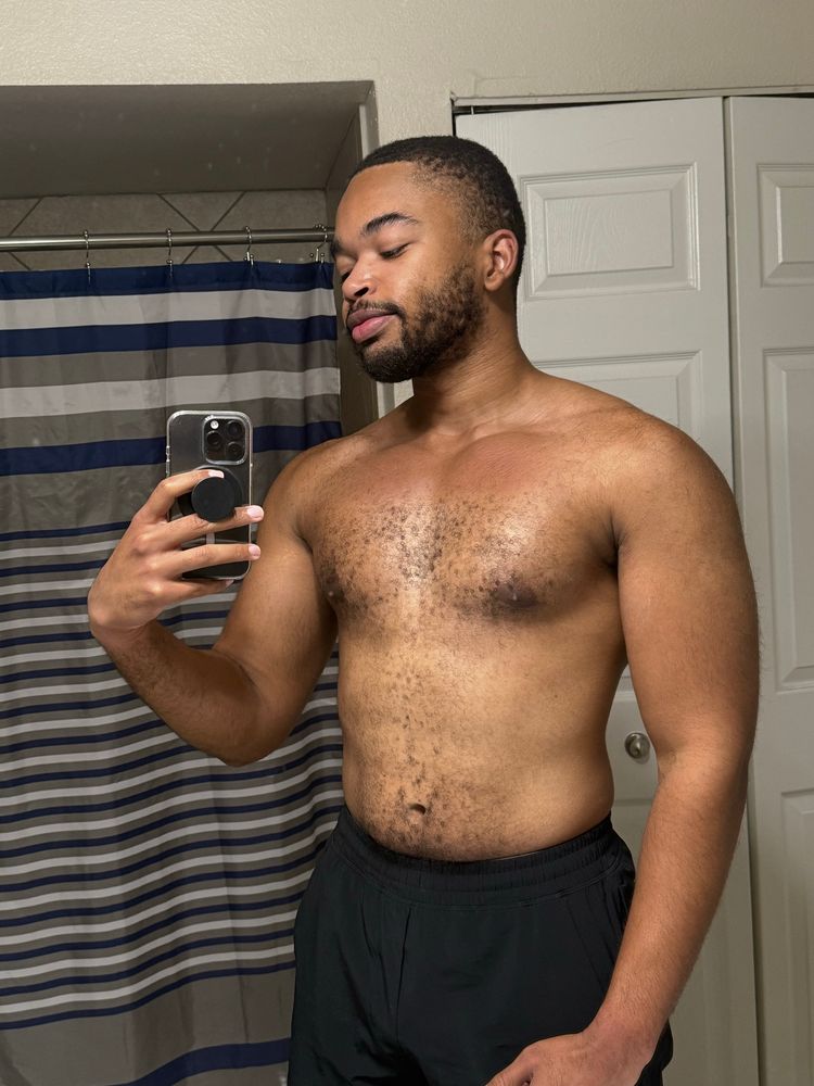 Shirtless mirror selfie of me looking at my phone as I take a post upper body workout pic in black shorts.