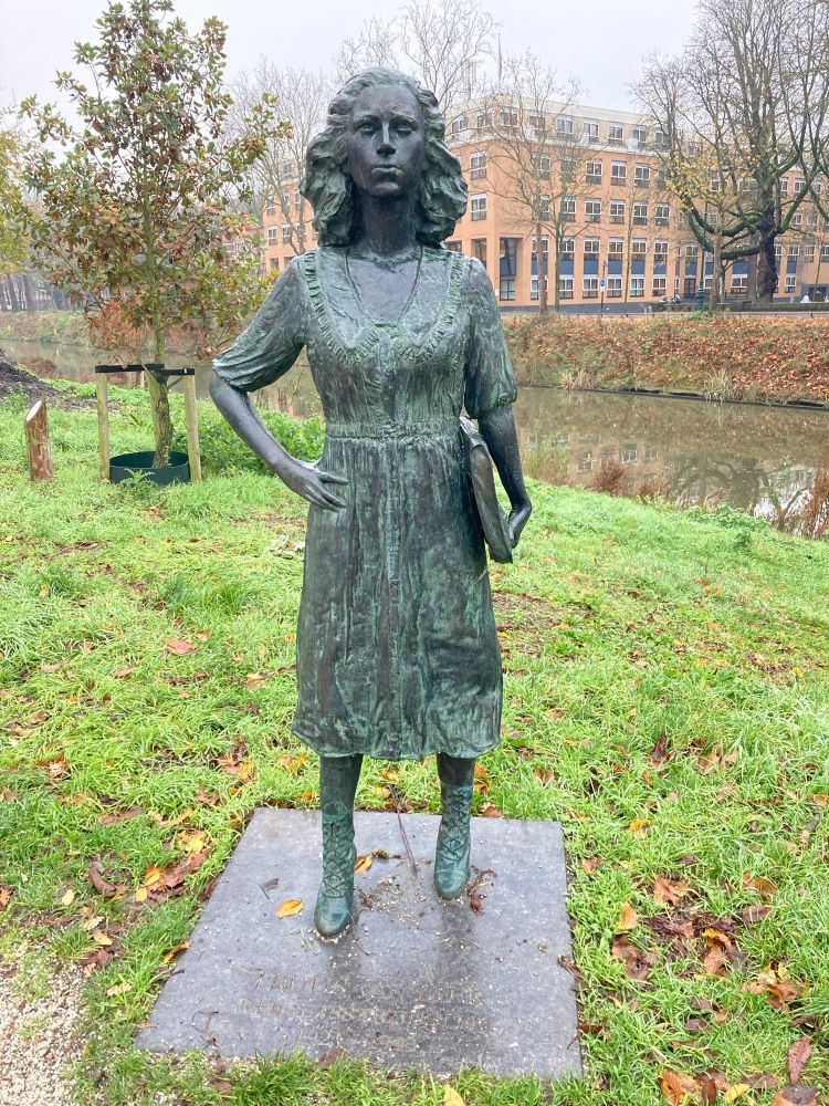 Een bronzen beeld van een vrouw in een jurk met een tas onder haar arm. Het beeld staat in een plantsoen langs het water. De bomen zijn bijna kaal.