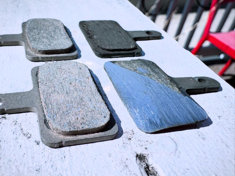 A pair of bike disc brake pads worn down past bare metal, next to a new fresh set of pads. 
