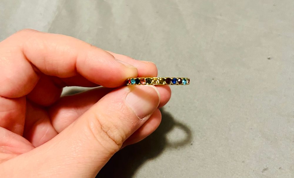 Close-up of a hand holding a gold ring. The ring is inset with small, multi-colored gemstones. The patterns shown is: pink, grey, brown, gold, dark red, dark blue, purple, light blue.