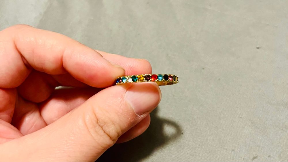 Close-up of a hand holding a gold ring. The ring is inset with small, multi-colored gemstones. The patterns shown is: green, yellow, purple, red, light blue, black, pink.