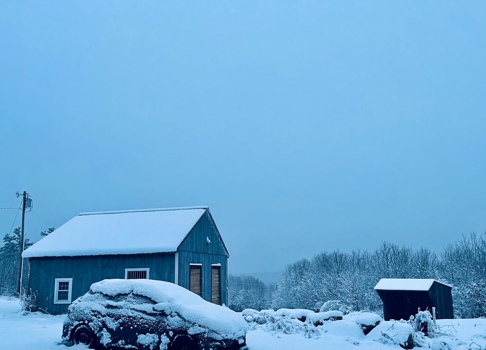 In the foreground is a black car covered in approximately two to three inches of snow. Behind the car is a blue barn with two large front doors; the roof also has a layer of snow. Separated by a small rock wall, a black shed on the side also has a layer of snow on it’s tilted roof. In the background, telephone wires are interspersed with trees laden with snow.