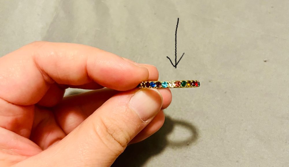 Close-up of a hand holding a gold ring. The ring is inset with small, multi-colored gemstones. The patterns shown is: light blue, white, pink, green. At green, the pattern begins to repeat.