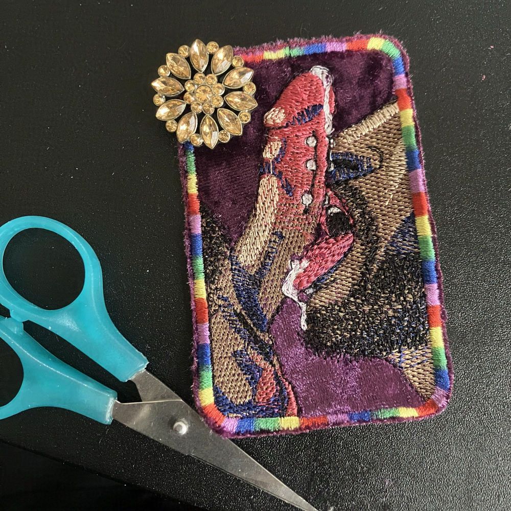 Photograph of an embroidered patch featuring an illustrated mouth and engorged phallus. A floral gem button is set atop the upper left corner of the patch. A small set of blue scissors is set next to the patch.