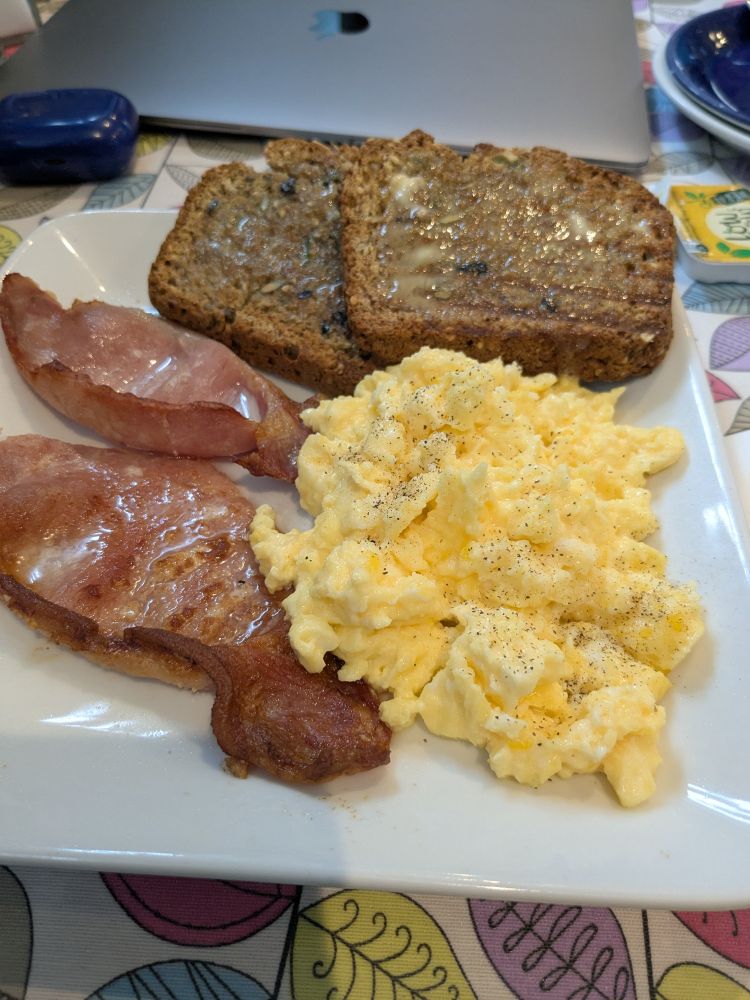 A square white plate with bacon, scrambled eggs and toasted home made brown bread. On the table behind them is a laptop and an earphone case.