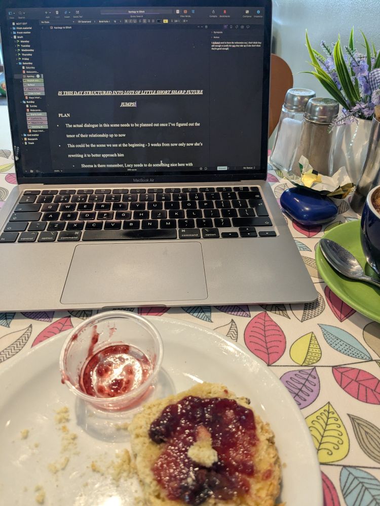 A laptop open on Scrivener, set to dark mode, with a half eaten scone and jam on a plate in front of it